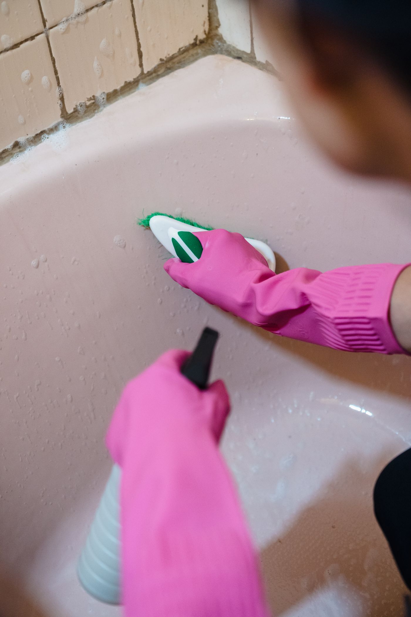 A person wearing pink gloves brushing a bathtub surface beside tiled walls.