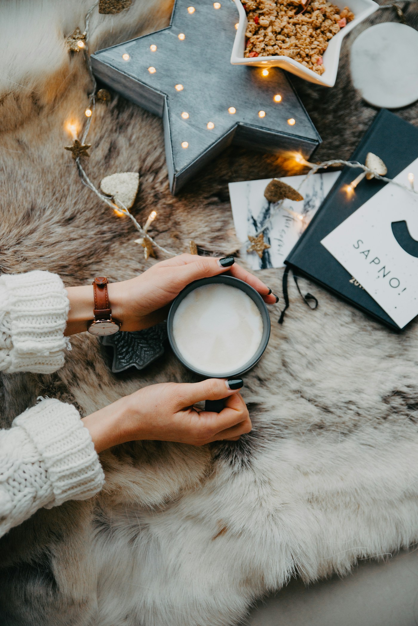 Hands holding a cup of a light-colored drink above a furry rug or textile surface with books and decorative objects nearby.