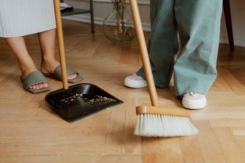 People using a broom and dustpan together on a wooden floor indoors.