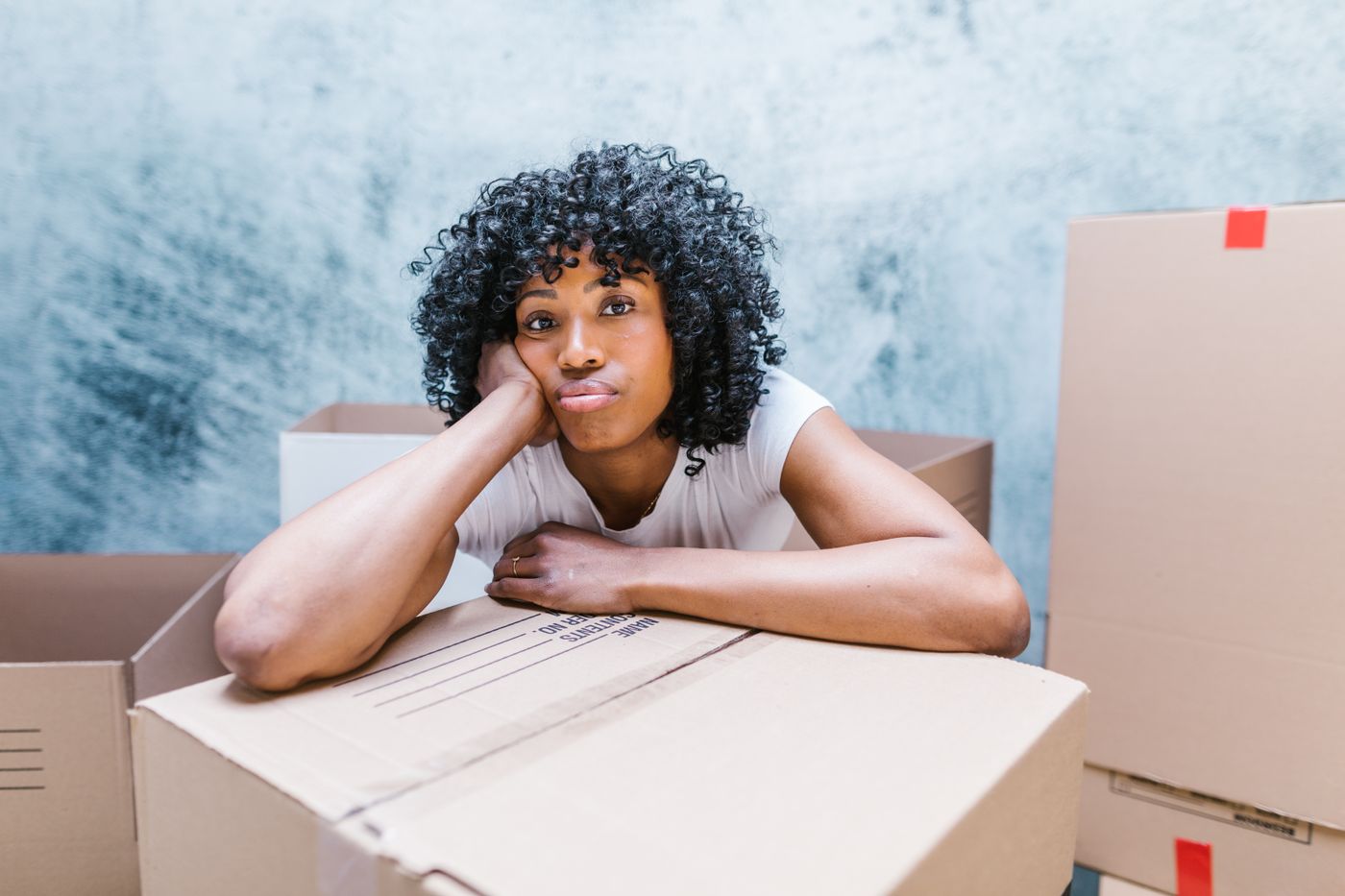 A woman resting her arms on a cardboard box among other moving boxes in an interior room.
