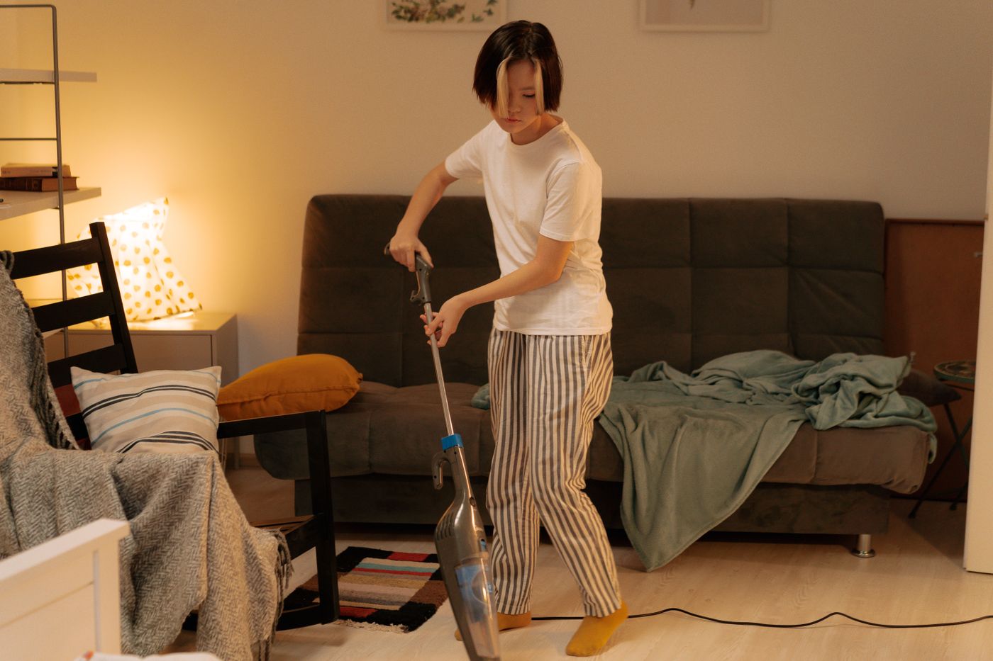 A woman using a stick vacuum beside a sofa, chair and small rug in a furnished room.