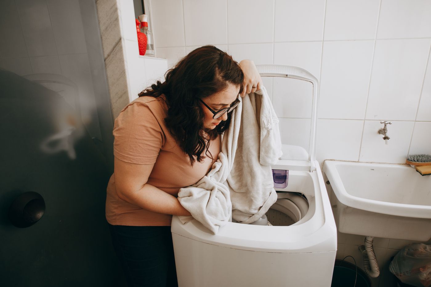 A woman handling laundry at a top-loading washing machine beside a utility sink.