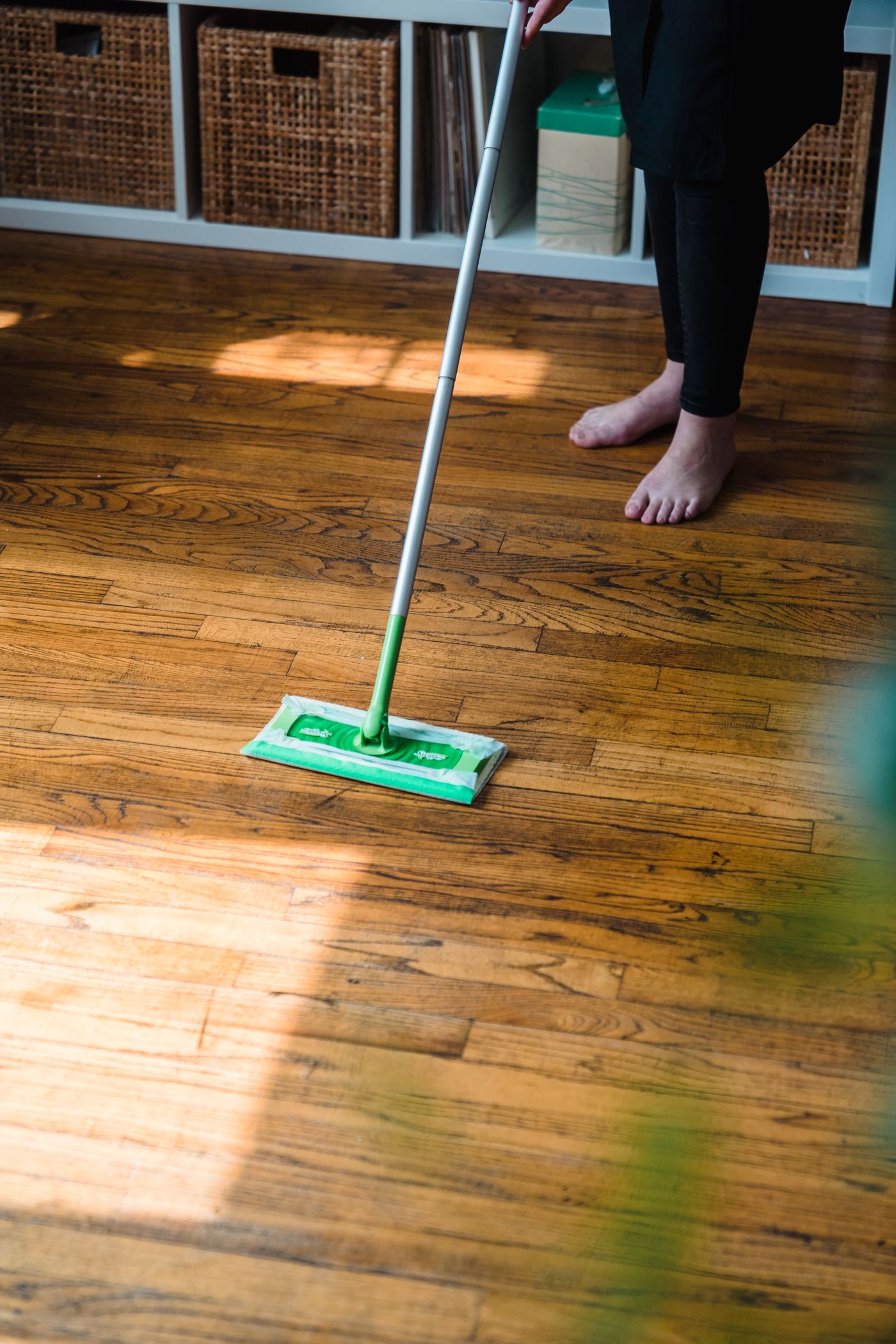 A person using a flat floor-cleaning tool on a wood-grain parquet-pattern floor beside storage furniture.